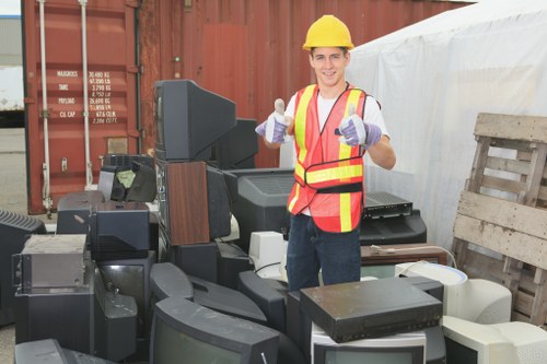 A young male worker wearing a yellow safety helmet, reflective orange vest with yellow stripes, and purple gloves standing in an outdoor area surrounded by various discarded electronic waste items, including outdated televisions and computer monitors with black and grey casings. The electronics are stacked in a cluttered manner on the pavement, with some partially leaning against a wooden pallet to the right and a shipping container in the background. The worker is smiling and giving a thumbs-up gesture, indicating a successful rubbish removal service. The scene is well-lit with natural daylight, emphasizing the textures of the electronic debris and the details of the worker's safety gear, illustrating the task of clearing unwanted electronic waste as part of a professional waste clearance operation in the Twickenham area near Feltham, managed by Feltham House Clearance.