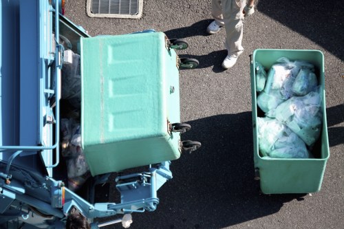 Aerial view of a waste collection scene showing two large, light green skip bins filled with assorted rubbish, located on a paved surface. One bin is positioned in front of a blue waste vehicle, which appears to be a skip loader or waste collection truck, with parts of its machinery and side visible. The second bin, filled with plastic bags and waste materials, is situated to the right of the vehicle. Part of a person wearing light-colored trousers and shoes is visible near the bins, indicating active rubbish removal. The scene suggests a typical urban or suburban environment, possibly in or near Feltham, with clean, well-maintained surroundings. This visual context reflects the services offered by Feltham House Clearance for rubbish removal and waste disposal, providing an accurate depiction of the process involved in clearing and collecting waste materials from residential or commercial properties.