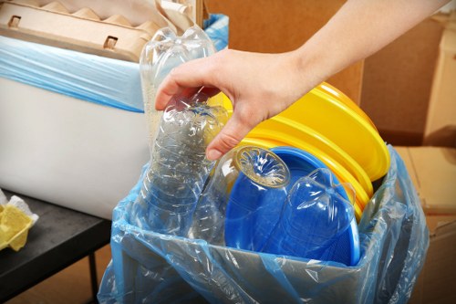 A close-up view of a person's hand gripping a clear plastic water bottle, which is partially crushed, as it is being placed into a large blue waste collection bag lined with a transparent plastic sheet. Inside the bag are several small, brightly coloured plastic buckets with yellow, blue, and orange finishes, and a rolled-up yellow item. The background shows a portion of a room with some cardboard boxes and a black surface, indicating a cluttered interior space likely prepared for rubbish clearance. The scene suggests activities related to decluttering or waste disposal, typical of rubbish removal services such as those offered by Feltham House Clearance, operating in the local Feltham area, TW postcodes, with an emphasis on environmentally mindful disposal of household waste.